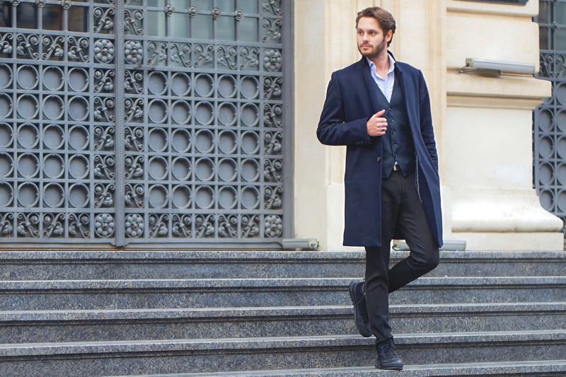 4 - A man wearing a classic navy blue overcoat, a tailored vest, a light blue dress shirt, and dark trousers while posing on a set of stone steps
